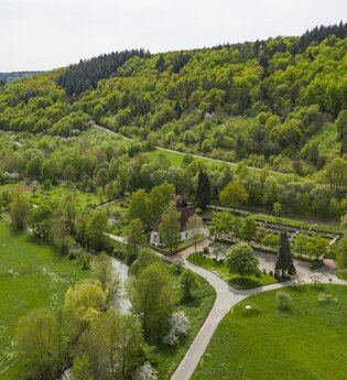 Luftbildaufnahem auf einen Parkplatz/Wohnmobilstellplatz bei Distelhausen. Daneben ist eine Kapelle und ein Friedhof zu sehen.  Es ist ein idyllischer Ort an der Tauber, mit Wiesen und bewaldeten Hügeln.