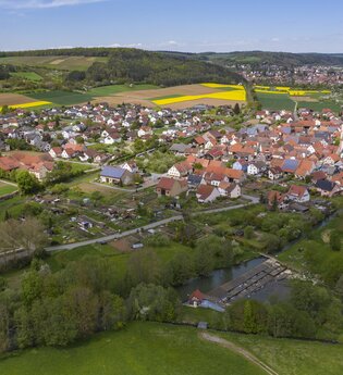 Luftbildaufnahme auf den Stadtteil Dittigheim, umrahmt von Wiesen, Feldern und Bäumen. Im Hintergrund ist die Stadt Tauberbischofsheim zu sehen.