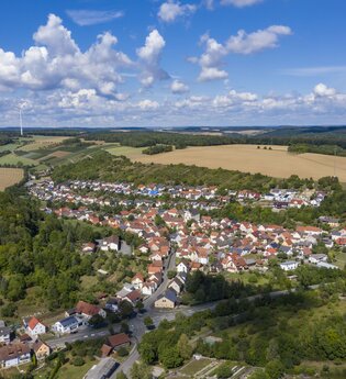 Luftbildaufnahme bei strahlendem Sonnenschein auf den Ort Dittwar. Der Ort liegt inmitten von Feldern und Wald. Im Hintergrund sind Windräder zu sehen.