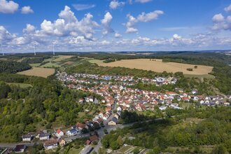 Luftbildaufnahme bei strahlendem Sonnenschein auf den Ort Dittwar. Der Ort liegt inmitten von Feldern und Wald. Im Hintergrund sind Windräder zu sehen.