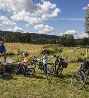 Zwei Erwachsene und zwei Kinder legen während einer Radtour eine Pause an der Tauber ein. Die Erwachsenen sitzen auf einer Holzbank. Der Bereich wurde renaturiert, Die Tauber ist im Hintergrund. zu sehen. Sie fließt im sanftem Bogen vorbei.