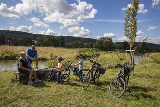 Zwei Erwachsene und zwei Kinder legen während einer Radtour eine Pause an der Tauber ein. Die Erwachsenen sitzen auf einer Holzbank. Der Bereich wurde renaturiert, Die Tauber ist im Hintergrund. zu sehen. Sie fließt im sanftem Bogen vorbei.