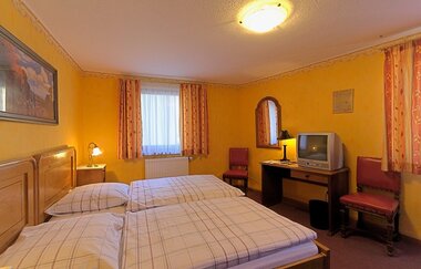 Das Zimmer ist gelb, es steht ein Doppelbett und ein Schreibtisch mit Stühlen im Zimmer