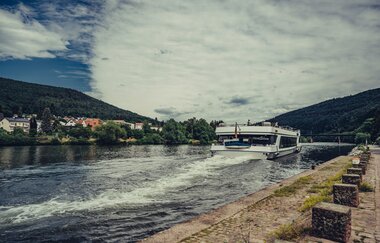 Ein weißess Ausflugsschiff welches auf einem Fluss fährt. Auf der linken Seite am Ufer sieht man eine Stadt. Neckargemünd im Odenwald.