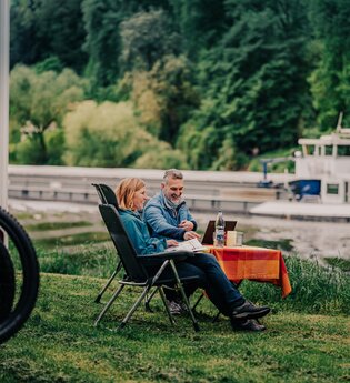 Ein Mann und eine Frau sitzen an einem Campingtisch vor Ihrem Wohnmobil am Ufer eines Flusses. Auf dem Fluß fährt ein Binnenschiff. Auf der anderen Uferseite ist Wald. Neckargemünd im Odenwald.