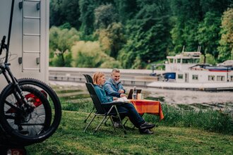 Ein Mann und eine Frau sitzen an einem Campingtisch vor Ihrem Wohnmobil am Ufer eines Flusses. Auf dem Fluß fährt ein Binnenschiff. Auf der anderen Uferseite ist Wald. Neckargemünd im Odenwald.