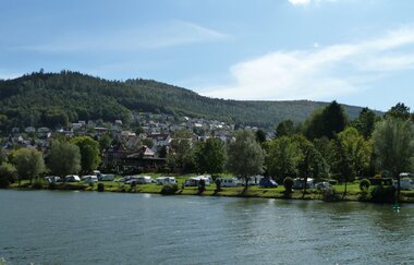 Im Vordergrund ist ein Fluß zu sehen (Neckar). Am Ufer des Flusses ist ein Campingplatz mit einer Reihe von Wohnwägen, mit einem bewaldeten Berg im Hintergrund. Eberbach im Odenwald.