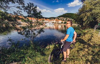 Ein Radfahrer mit blauem T-Shirt steht mit seinem Rad an einem Flussufer. Auf de anderen Uferseite sieht man eine Stadt. Eberbach im Odenwald.