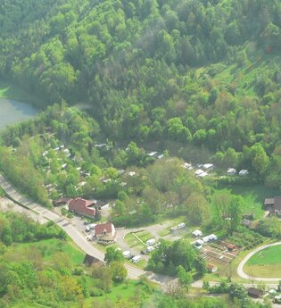 Luftbildaufnahme auf den im Tal gelegenen und mit vielen Bäumen bepflanzten Campingplatz Romantische Strasse in Creglingen-Münster. Der Hang ist bewaldet und im Hintergrund ist ein kleiner See zu sehen.