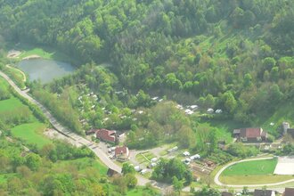 Luftbildaufnahme auf den im Tal gelegenen und mit vielen Bäumen bepflanzten Campingplatz Romantische Strasse in Creglingen-Münster. Der Hang ist bewaldet und im Hintergrund ist ein kleiner See zu sehen.