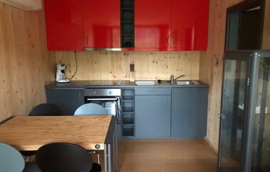 Eine Küchenzeile in rot und grau, ein Tisch mit Stühlen und ein Kühlschrank stehen im Wohnraum