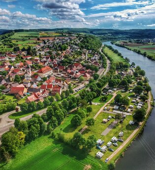 Drohnenaufnahme mit Blick auf einen Fluss (Neckar). Auf der rechten Uferseite sieht man einen Campingplatz und dahinter einen angrenzenden Ort. Binau im Odenwald.