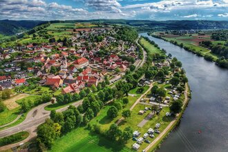 Drohnenaufnahme mit Blick auf einen Fluss (Neckar). Auf der rechten Uferseite sieht man einen Campingplatz und dahinter einen angrenzenden Ort. Binau im Odenwald.