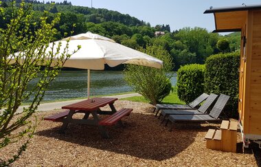 Ein Fluss dahienr eine Sitzgruppe aus Hol mit einem weissen Sonnenschrift. Dahinter stehen zwei Sonnenliegen mit einem Schäferwagen. Binau im Odenwald