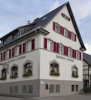Der Gasthof von außen hat rote Fensterläden und ist weiß und unten grau gestrichen