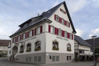 Der Gasthof von außen hat rote Fensterläden und ist weiß und unten grau gestrichen