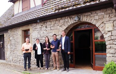 Drei Männer und zwei Frauen stehen mit einem Glas Wein vor dem Eingang vom Weingut, Besenwirtschaft und Kelterhotel