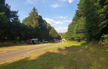 Auf dem Bild ist der Naturcampingplatz in Bad Mergentheim zu sehen, ein Weg mit Wiesenflächen rechts und links und  großen Bäume im Hintergrund. Ein Stellplatz ist mit Zelt/Auto und Motorrad belegt. Ein sonniger Tag mit blauem Himmel und weißen Wolken..