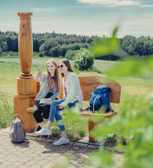 2 Frauen sitzen auf einer Holzbank mit einer Holzsäule und machen ein Selfie. Im Hintergrund sind Wald und Wiesen. Odenwald.