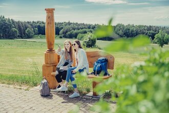 2 Frauen sitzen auf einer Holzbank mit einer Holzsäule und machen ein Selfie. Im Hintergrund sind Wald und Wiesen. Odenwald.