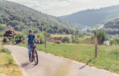 Ein Radfahrer mit blauem Shirt fährt einen Radweg hoch. Im Tald ist ein Fluß mit bergigen und bewaldeten Ufern. Odenwald.
