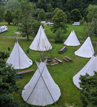 Blick von oben auf eine Wiese mit 6 Tipis die in einem Kreis stehen. In der Mitte ist eine Feuerstelle mit Bänken. Neckargerach im Odenwald.