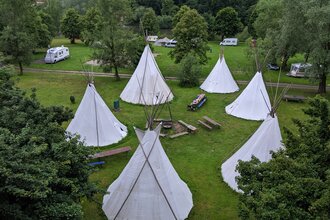 Blick von oben auf eine Wiese mit 6 Tipis die in einem Kreis stehen. In der Mitte ist eine Feuerstelle mit Bänken. Neckargerach im Odenwald.