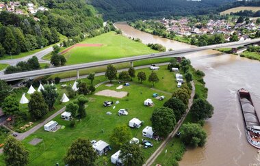 Ein Fluss mit einem Binnenschiff und einer großen Straßenbrücke die über den Fluß geht. Auf der rechten Uferseite ist eine Stadt zu sehen. Auf der linken Seite en Campingplatz mit Zelten und Wohnmobilen. Neckargerach im Odenwald