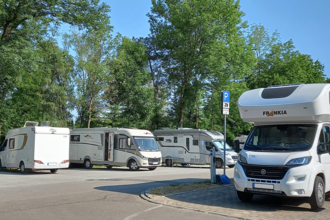 Wohnmobilstellplatz mit mehreren geparkten Wohnmobilen und Bäumen im Hintergrund.