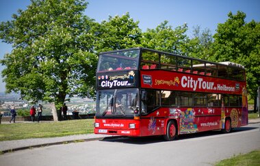 der rote Doppeldeckerbus mit Cabriodach fährt auf einer asphaltierten Straße. Die Sonne scheint, im Hintergrund sind grüne, hohe Bäume erkennbar
