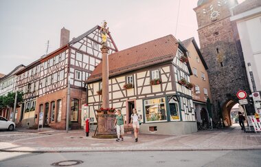 Zwei Fachwerkhäuser mit einer Sandsteinsäule im Vordergrund auf der eine goldene Figur ist. 2 Frauen die an der Säule vorbeilaufen. Neben den Fachwerkhäusern ist ein Stadtturm mmit Uhr. Buchen im Odenwald
