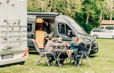 Ein weisses und ein graues Wohnmobil. 2 Männer und 2 Frauen die an einem Campingtisch sitzen und sich zuprosten. Buchen im Odenwald