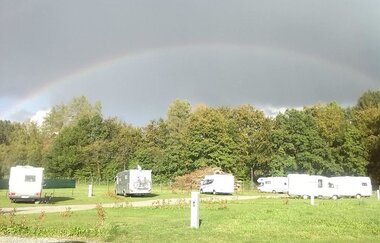 Begrünter Campingplatz mit Wohnmobilen und Regenbogen am Himmel
