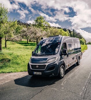Dunkler Kastenwagen fährt auf einer Straße. Links vom Wohnmobil ist eine Streuobstwiese.| Odenwald