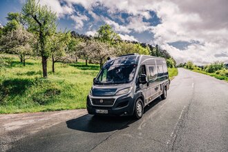 Dunkler Kastenwagen fährt auf einer Straße. Links vom Wohnmobil ist eine Streuobstwiese.| Odenwald