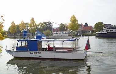 Ein Flug mit einem kleineren weiß-blauen Motorboot mit einer Wimpelkette. Im Hintergrund ein Dorf mit Bäumen. Haßmersheim im Odenwald.