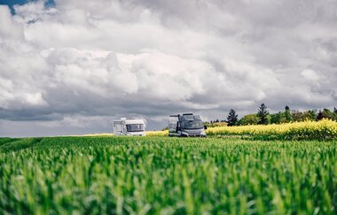 Ein grüne Wiese, dahinter fahren ein weißes und ein graues Wohnmobil. Im Hintergrund ist ein gelbes Rapsfeld. Odenwald.