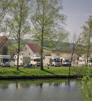 Blick auf den direkt an der Tauber gelegenen Wohnmobilstellplatz in Röttingen. Der Platz ist gut besucht. Im Hintergrund sind Häuser zu sehen.
