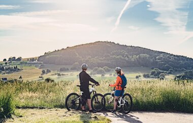Zwei Radfahrer stehen vor einer Wiese und schauen auf eine Erhebung. Waldbrunn im Odenwald.