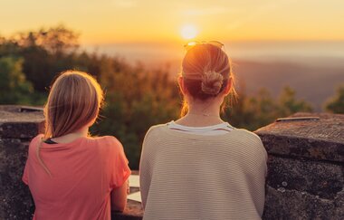 2 Frauen stehen am Rand eines Turms und schauen in die Weite und den Sonnenuntergang. Katzenbuckel im Odenwald