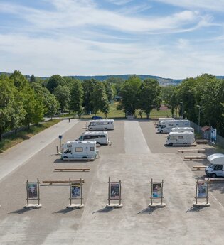 Luftaufnahme vom Stellplatz. Im Vordergrund wird der Platz durch Begrüßungstafeln abgegrenzt, Wohnmobile stehen auf dem Schotterplatz.