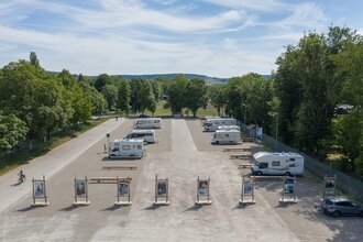 Luftaufnahme vom Stellplatz. Im Vordergrund wird der Platz durch Begrüßungstafeln abgegrenzt, Wohnmobile stehen auf dem Schotterplatz.