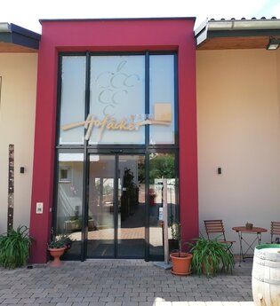 Blick auf den Eingangsbereich mit Glasfront vom Weingut Hofäcker in Weikersheim-Queckbronn. Im Hof davor ist ein Sitzgruppe sowie Blumen und Weinfässer als Dekoration zu sehen.