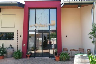 Blick auf den Eingangsbereich mit Glasfront vom Weingut Hofäcker in Weikersheim-Queckbronn. Im Hof davor ist ein Sitzgruppe sowie Blumen und Weinfässer als Dekoration zu sehen.