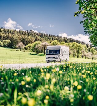 Weißes Wohnmobil fährt auf einer Strasse. Im Vordergrund ist eine Blumenwiese im Hintergrund eine Steuobstwiese mit angrenzendem Wald. | Odenwald