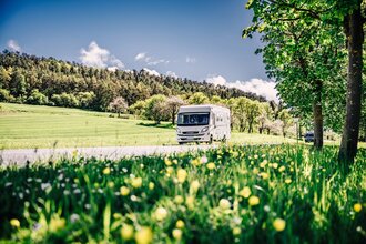 Weißes Wohnmobil fährt auf einer Strasse. Im Vordergrund ist eine Blumenwiese im Hintergrund eine Steuobstwiese mit angrenzendem Wald. | Odenwald