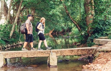 Ein Bach mit einer kleinen Steinbrücke. auf dieser laufen eine Frau und ein Mann. Im Hintergrund sind die Uferseiten stark bewachsen. Elztal im Odenwald