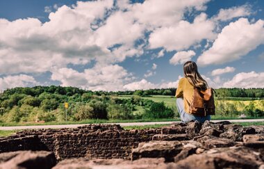Eine Frau mit gelbem Pullover sitzt auf den Mauern eines Römerkastells und blickt auf eine Landschaft mit Bäumen und Wiesen. Elztal im Odenwald.