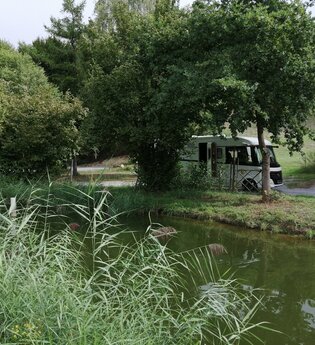 Ein Fischteich mit einem weißen Wohnmobil unter Bäumen. Elztal im Odenwald