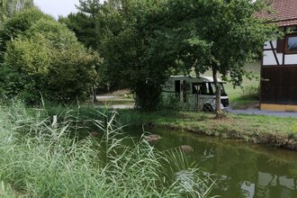 Ein Fischteich mit einem weißen Wohnmobil unter Bäumen. Elztal im Odenwald